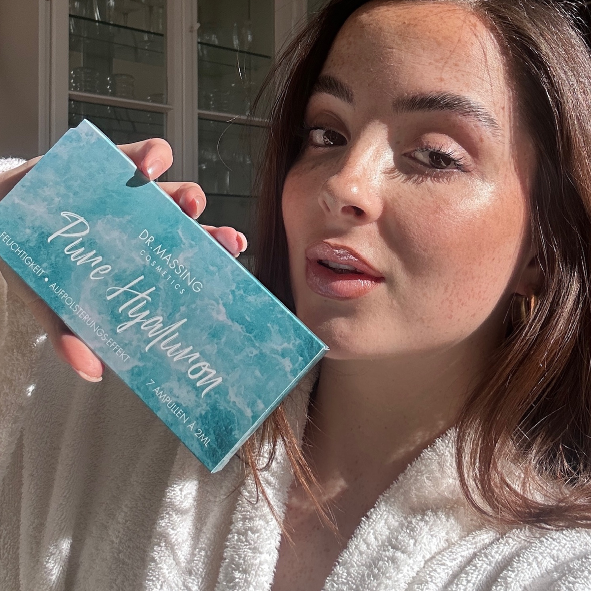 Frau hält Pure Hyaluron Ampullen Box in der Hand neben ihr Gesicht und schaut in die Kamera