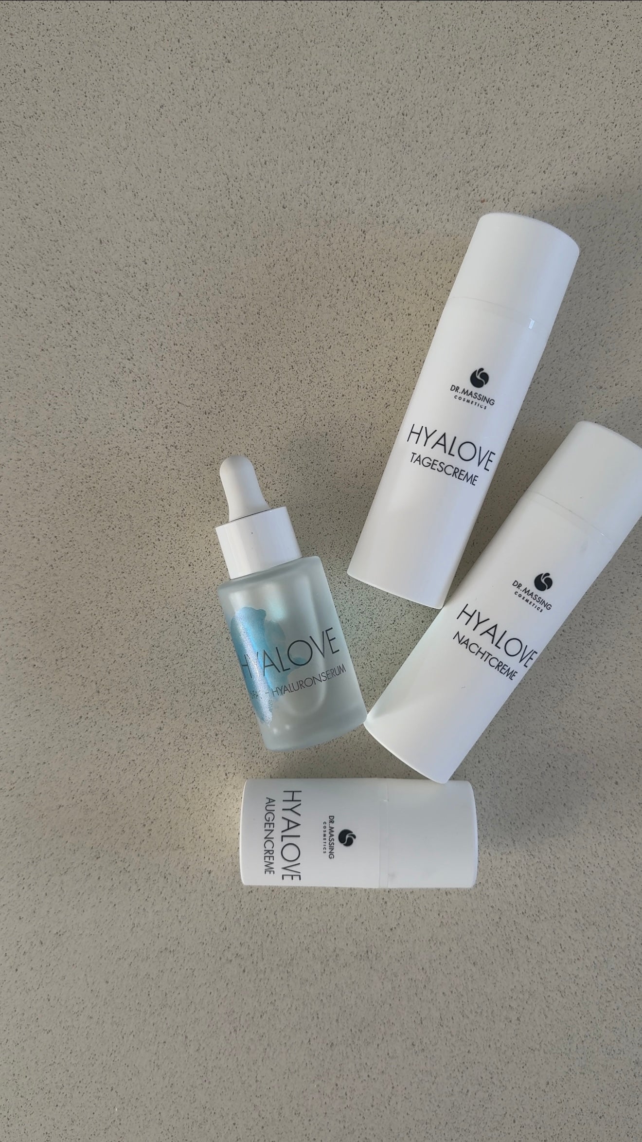 4er Set Hyalove Glass Skin Produkte mit Hyaluronserum, Tagescreme, Augencreme und Nachtcreme liegen auf sandfarbenen Tisch