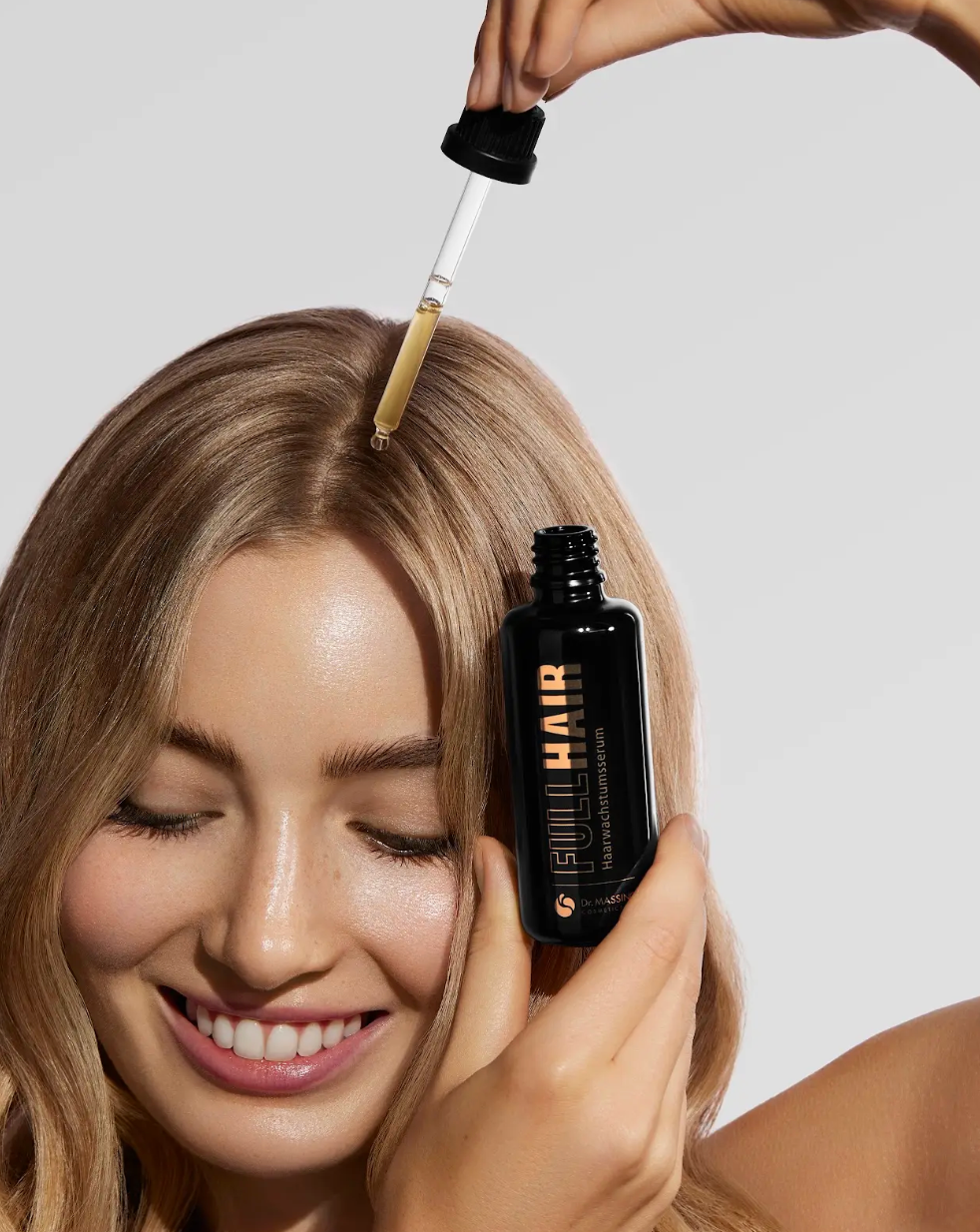 Frau trägt Haarwuchsmittel Serum mithilfe der Pipette direkt auf die Kopfhaut auf und lächelt dabei freundlich