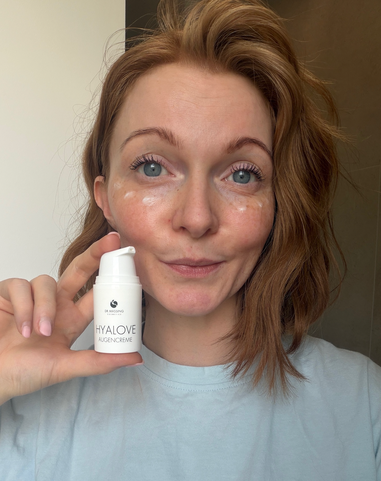 2. Schritt Anwendung Hyalove Augencreme mit Creme unter den Augen einer Frau die in die Kamera schaut und das Produkt neben ihrem Gesicht hält