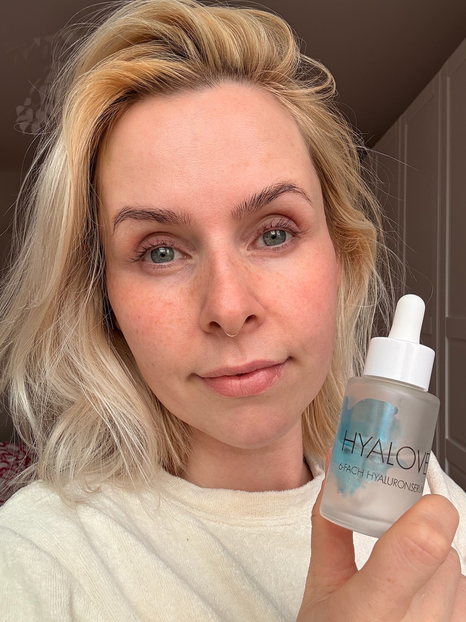 Junge Frau mittleren Alters mit schöner und gepflegter Haut hält Hyalove Serum neben ihr Gesicht und schaut glücklich in die Kamera