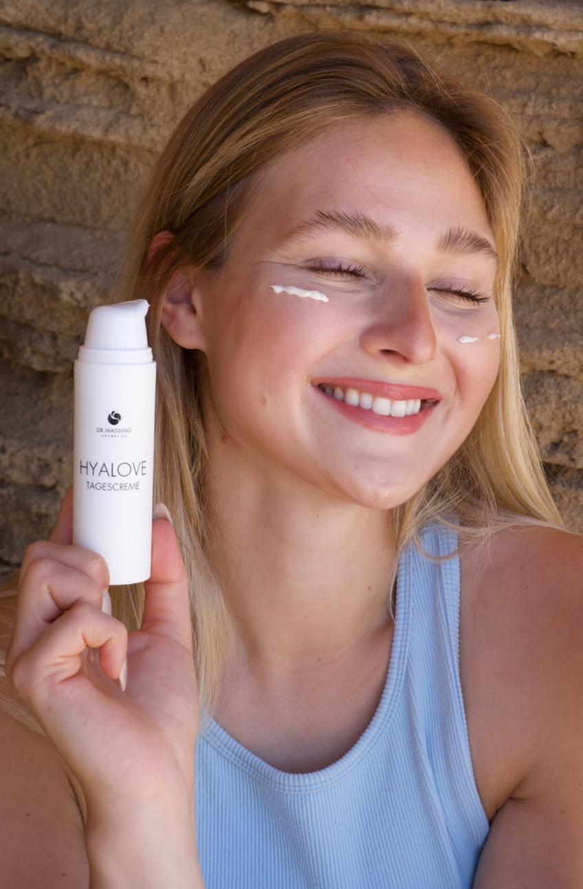 Portrait einer Frau mit geschlossenen Augen und lächelnden Ausdruck, die Hyalove Creme auf das Gesicht aufgetragen hat und Produkt neben ihr Gesicht hält