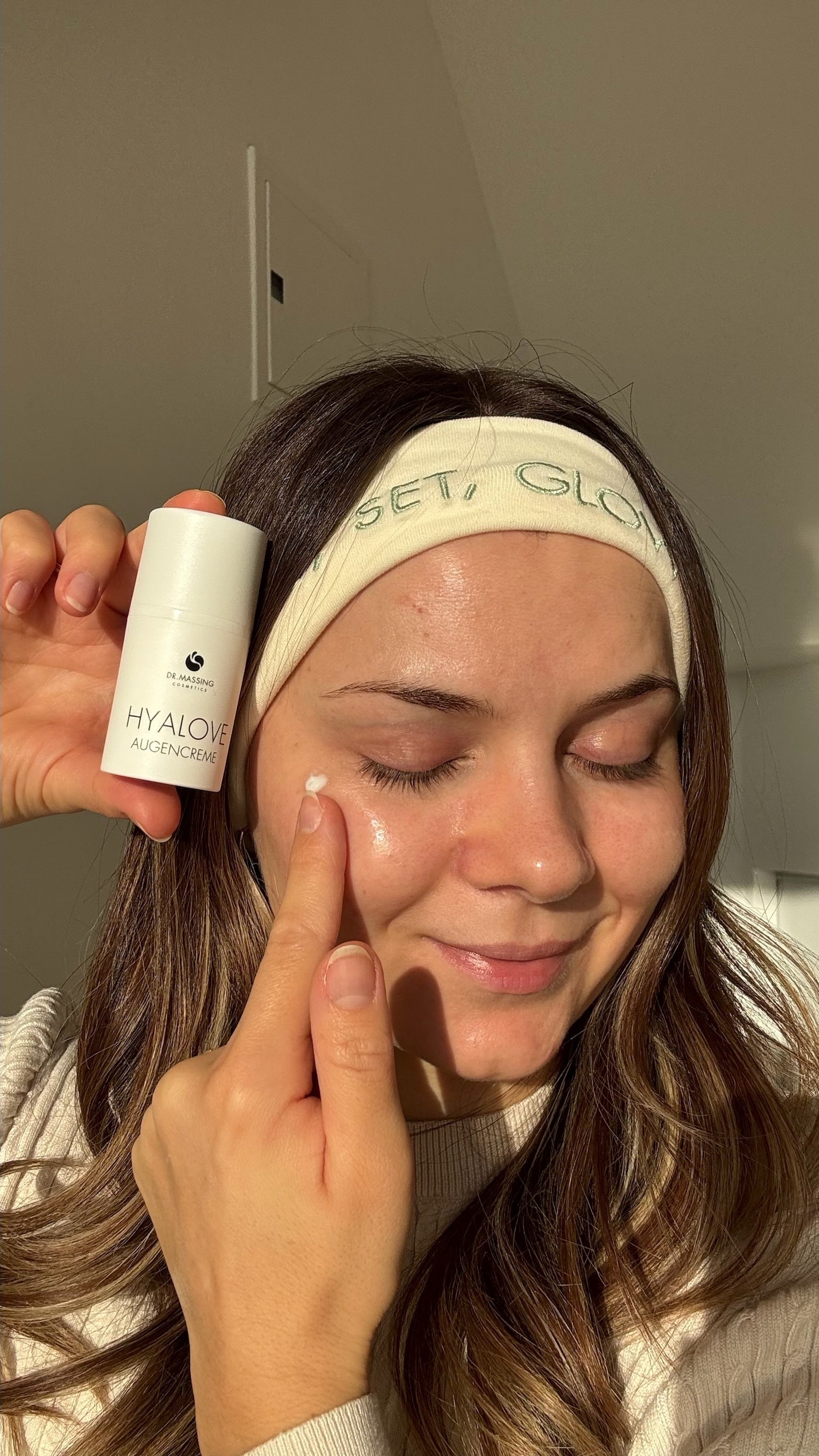 Frau sitzt mit Beauty Stirnband in der Sonne und trägt Augencreme von Dr. Massing auf und hält das Produkt neben ihr Gesicht