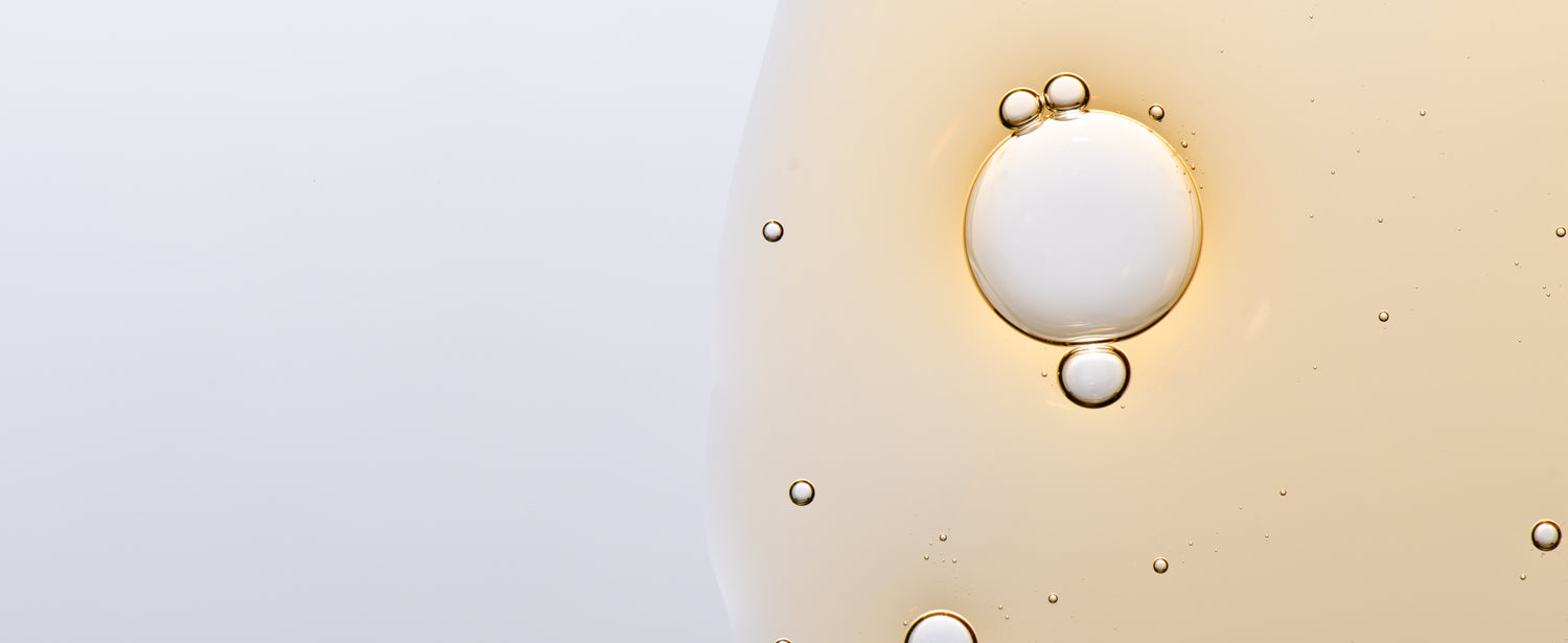 Eine Nahaufnahme eines Tropfens des Full Brow Augenserums, helle goldene Flüssigkeit mit eingeschlossenen Luftbläschen