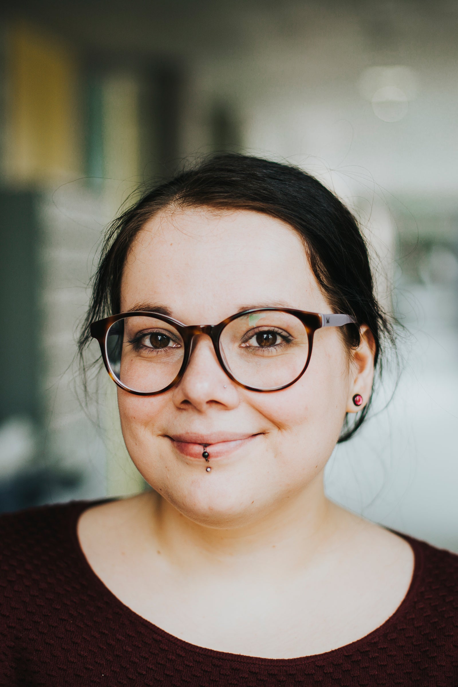 Portrait einer Frau mit junger gepflegter Gesichtshaut. Die Frau Trägt eine Brille und Piercings und lächelt freundlich in die Kamera.