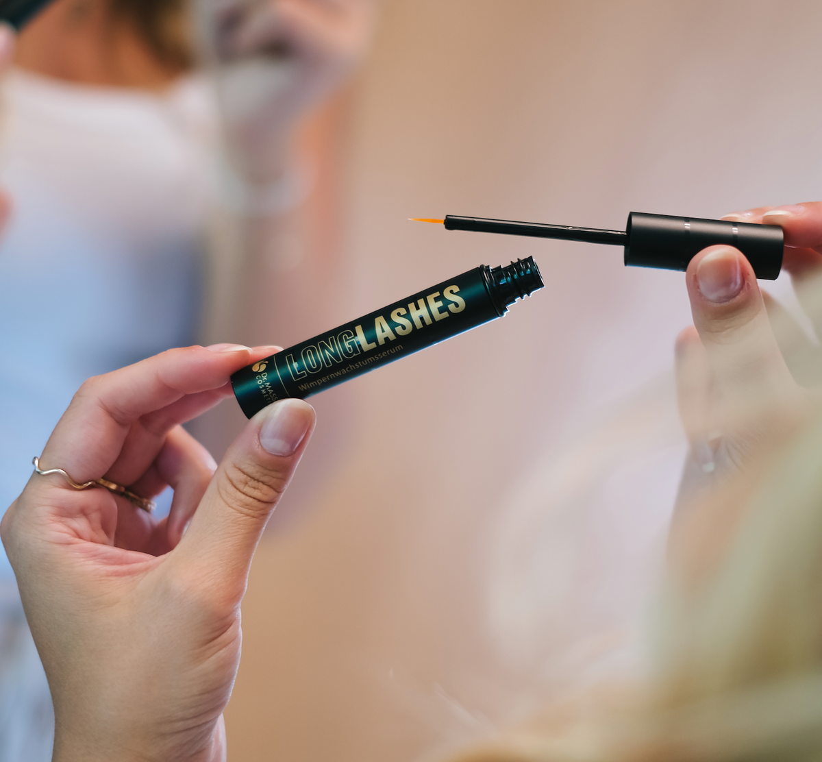 Zwei Hände welche Long Lashes Classic halten und den Pinsel aus Flasche für Anwendung Schritt 1 herausziehen